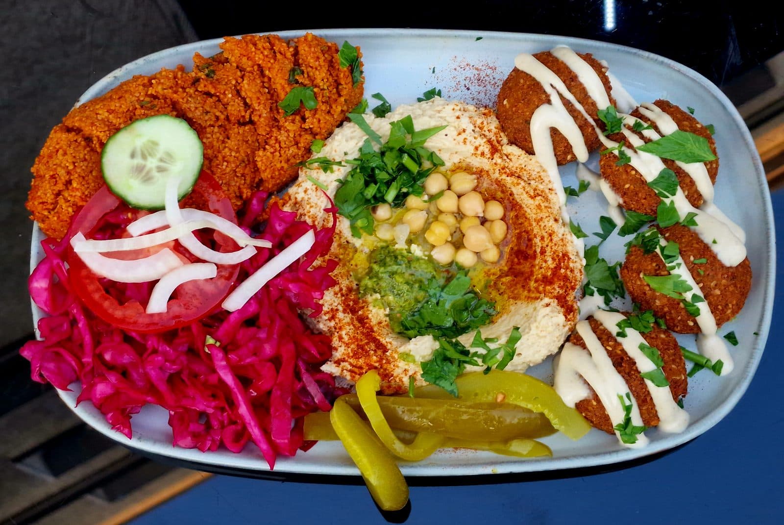 Falafel with tahini drizzle beside hummus with chickpeas, pickled cabbage, pickles, tomato and bulgur salad, top-down