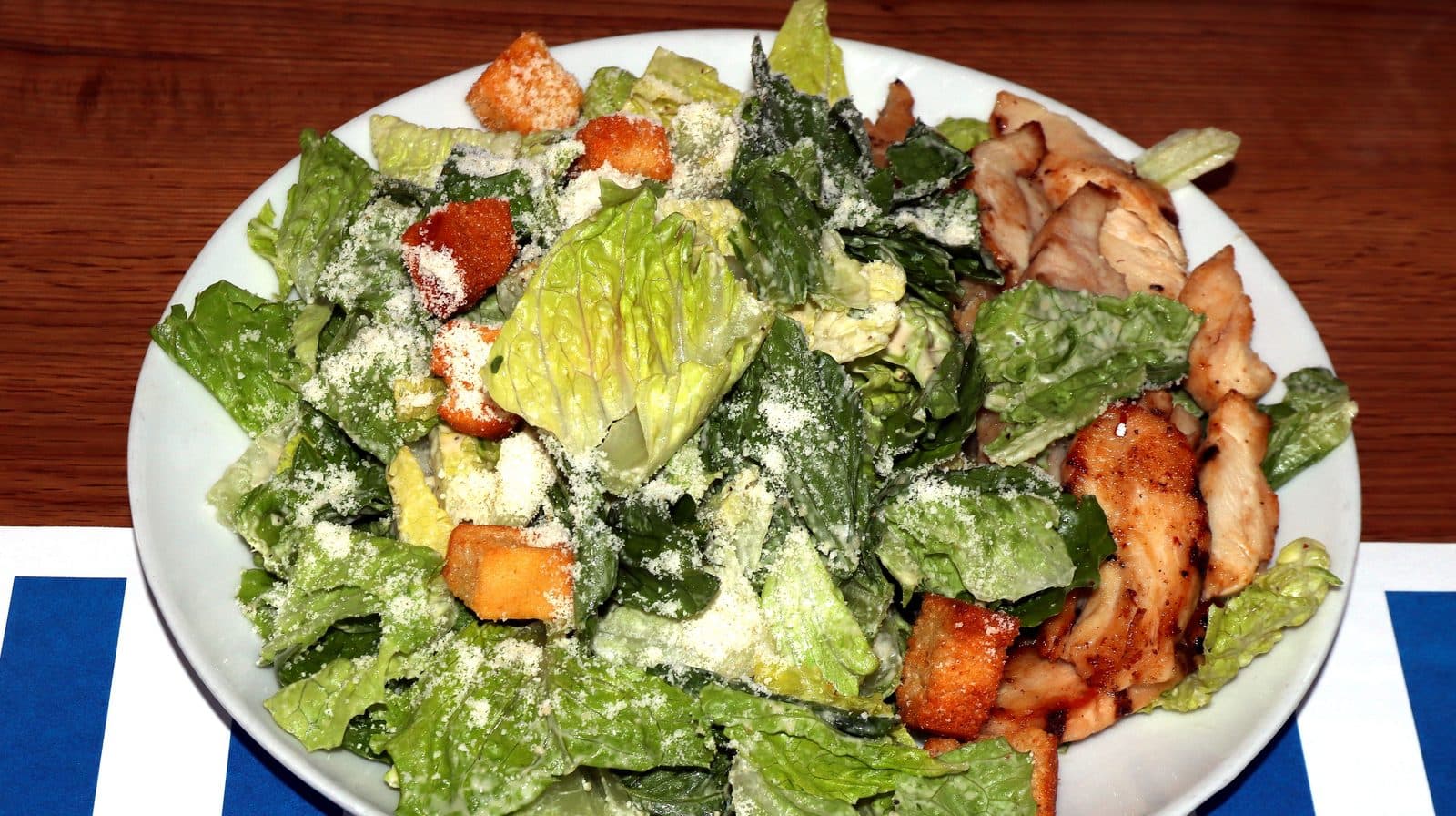 Caesar salad of chopped romaine, croutons and grated parmesan with sliced grilled chicken on a white plate