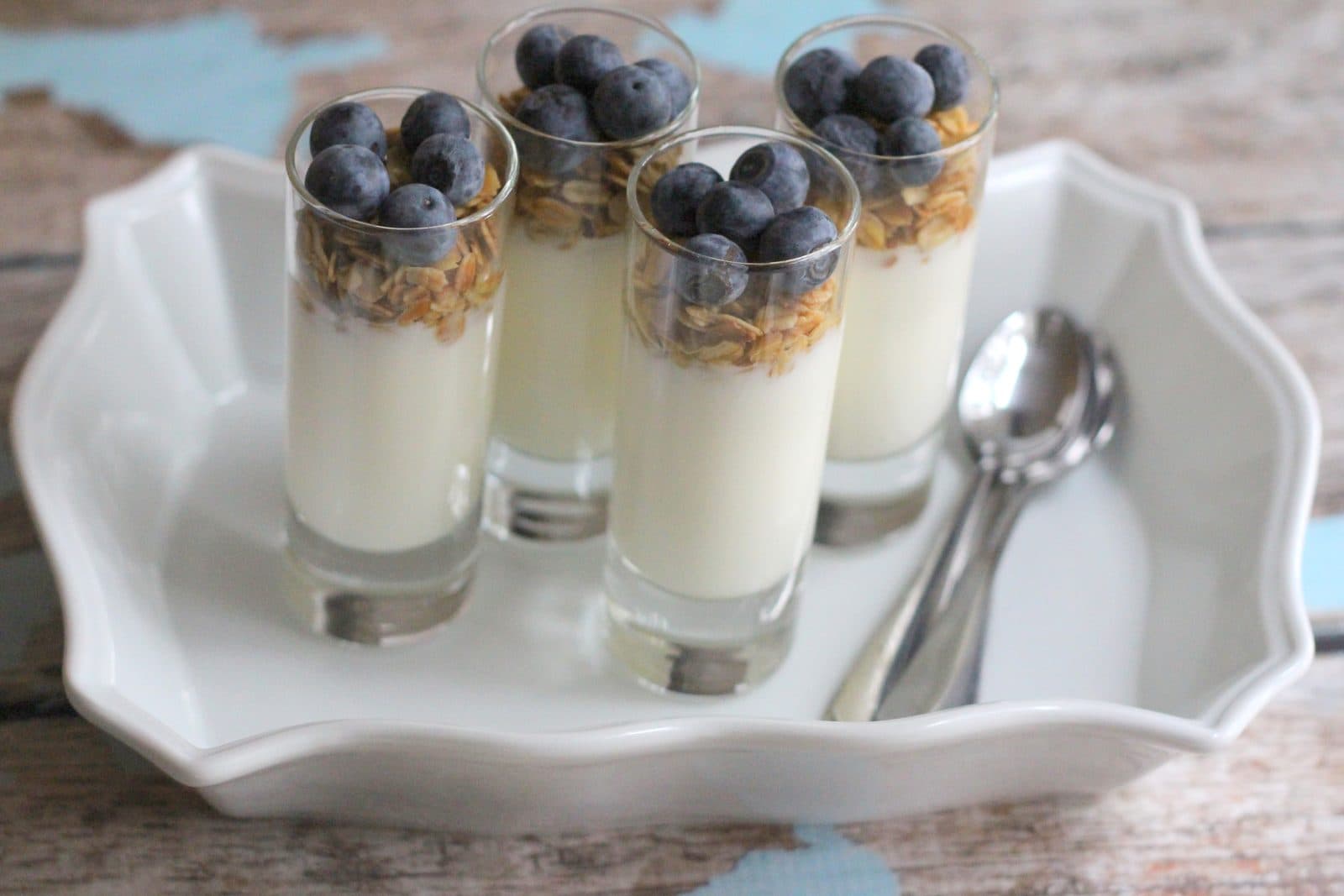 Four shot-glass yogurt parfaits topped with granola and blueberries on a white dish