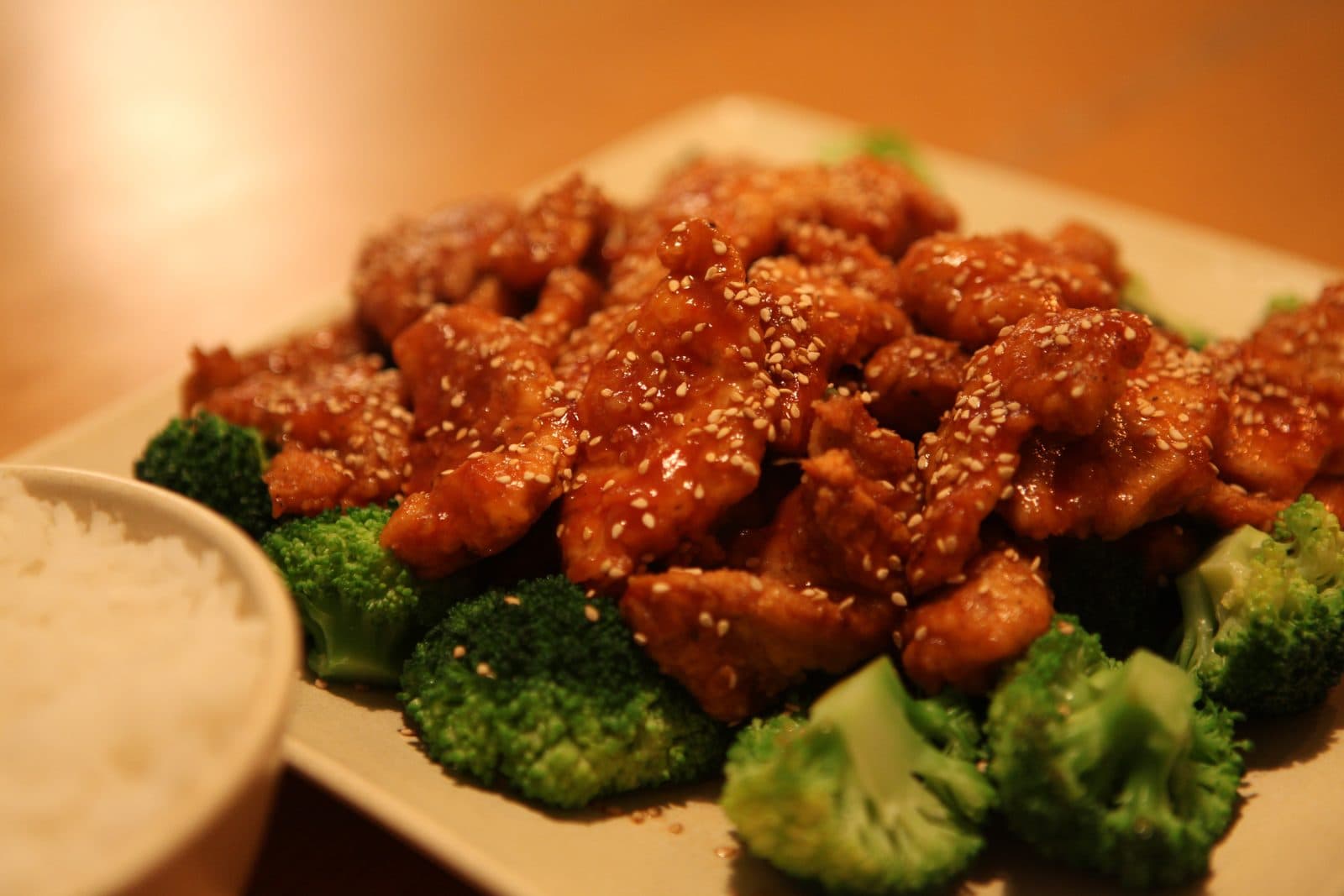 Sticky glazed chicken pieces with sesame seeds over broccoli, with a bowl of white rice