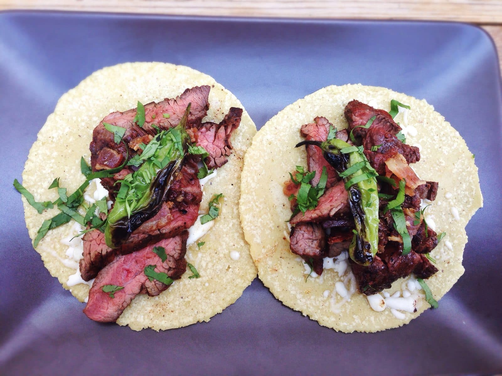 Two corn tortilla tacos topped with sliced grilled steak, a blistered green chilli and chopped coriander