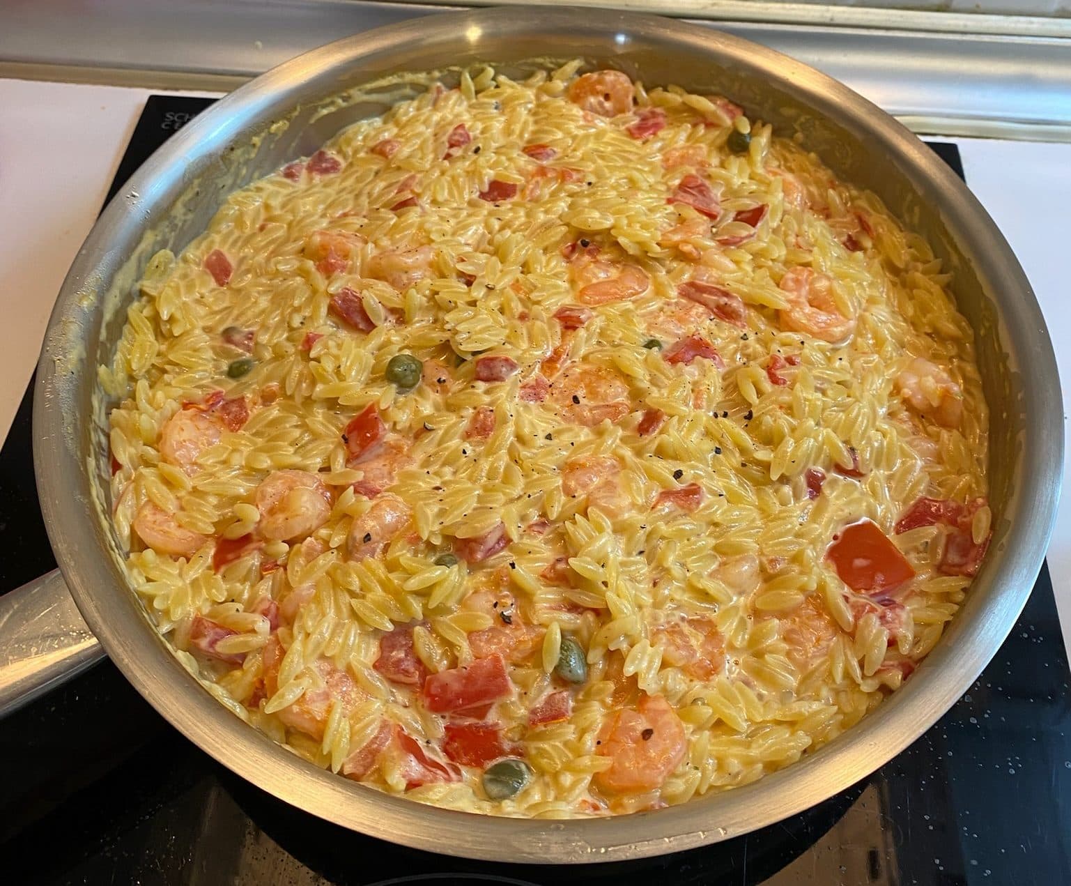 Creamy orzo with prawns, red pepper and capers in a stainless-steel pan on the hob