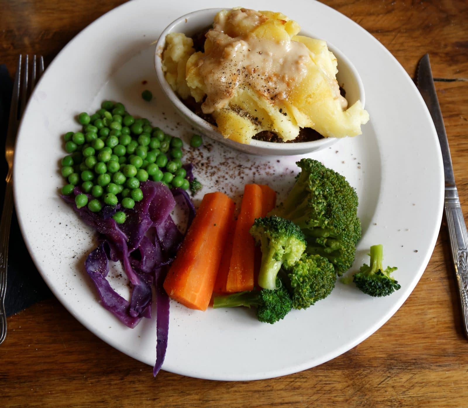 Cottage pie in an individual dish with peas, carrots, broccoli and red cabbage on a pub plate