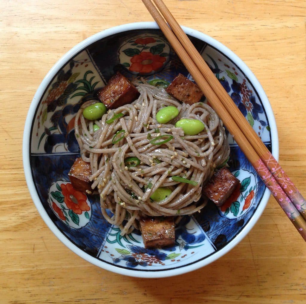 Soba noodles with browned tofu cubes, edamame, scallions and sesame in a blue patterned bowl with chopsticks, top-down