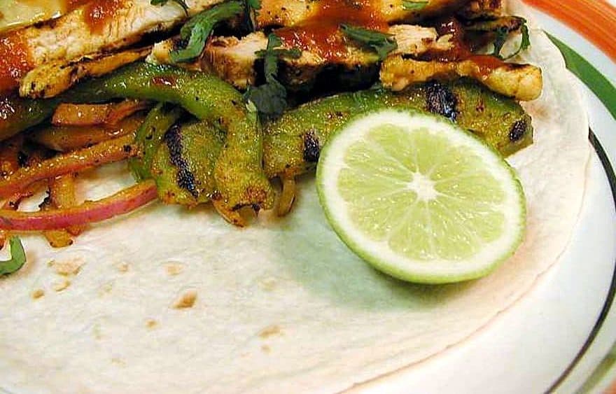 Grilled chicken strips with charred green peppers and red onion on a flour tortilla with a lime half