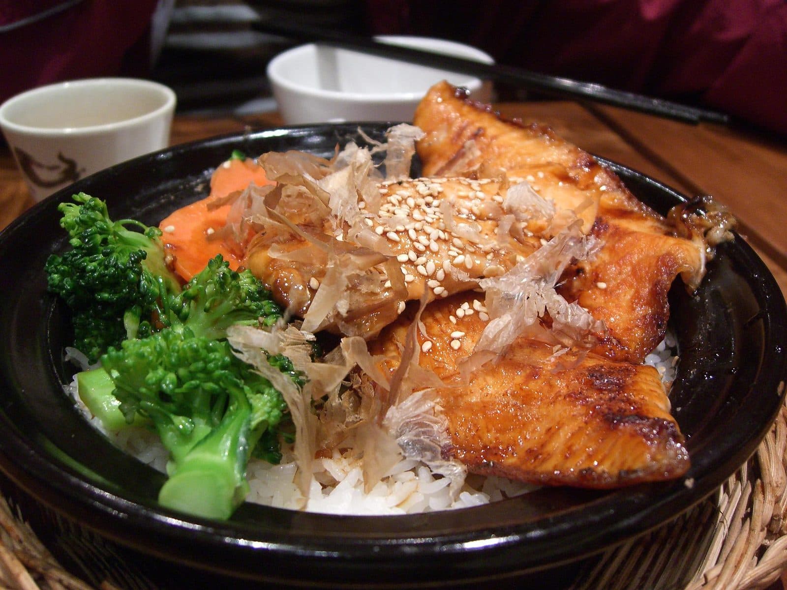 Glazed teriyaki salmon over rice with broccoli, carrot, sesame and bonito flakes in a black bowl