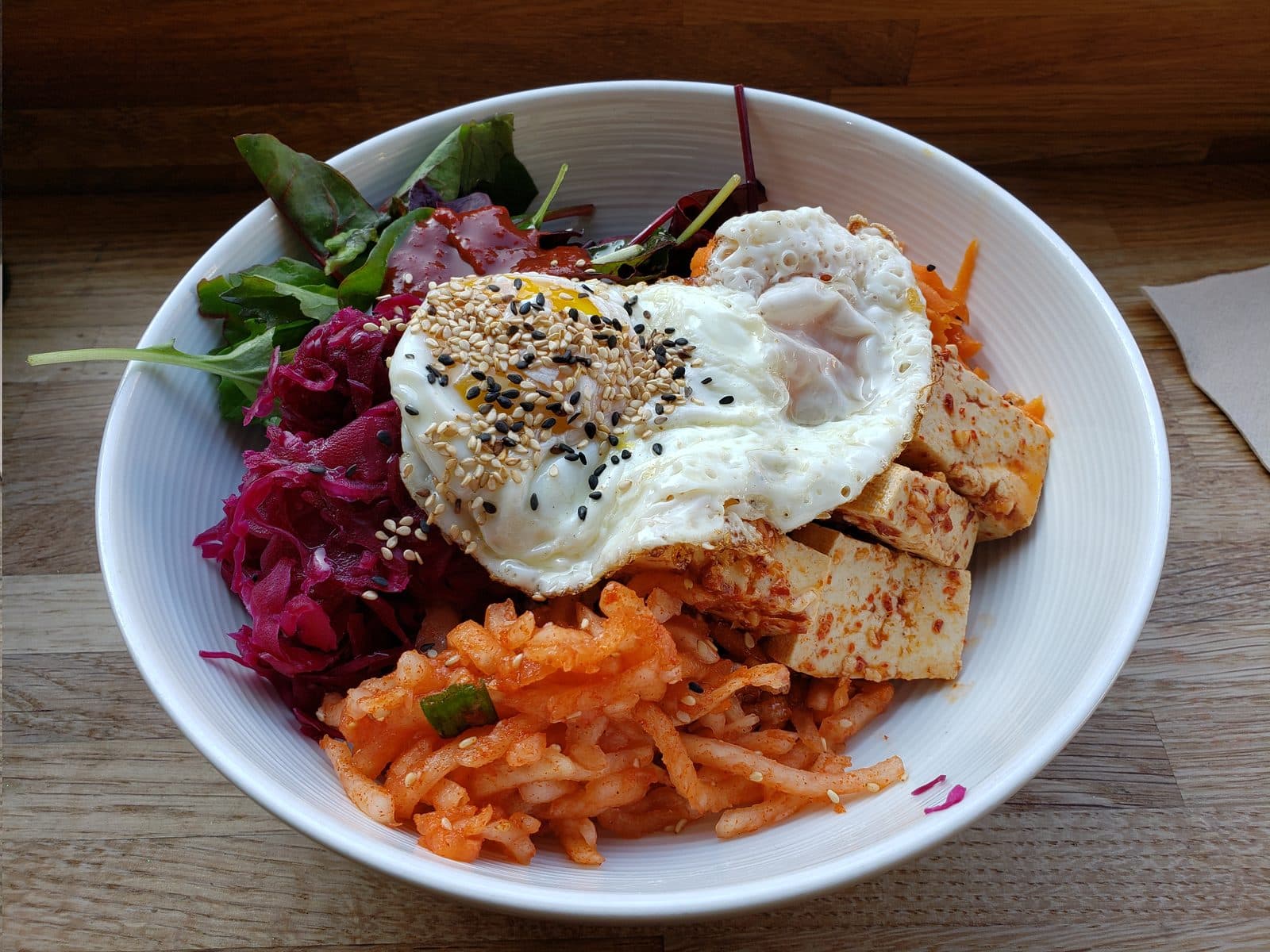 Tofu bibimbap bowl with a fried egg, kimchi radish, pickled red cabbage, carrot and greens