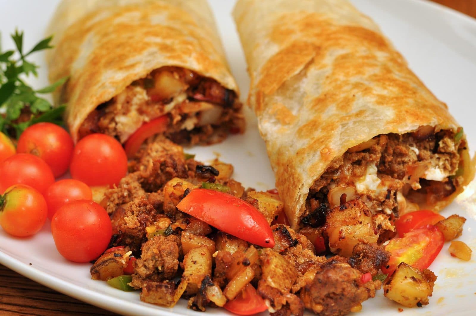 Two toasted breakfast burritos cut open to show sausage, egg, potato and pepper filling, with cherry tomatoes